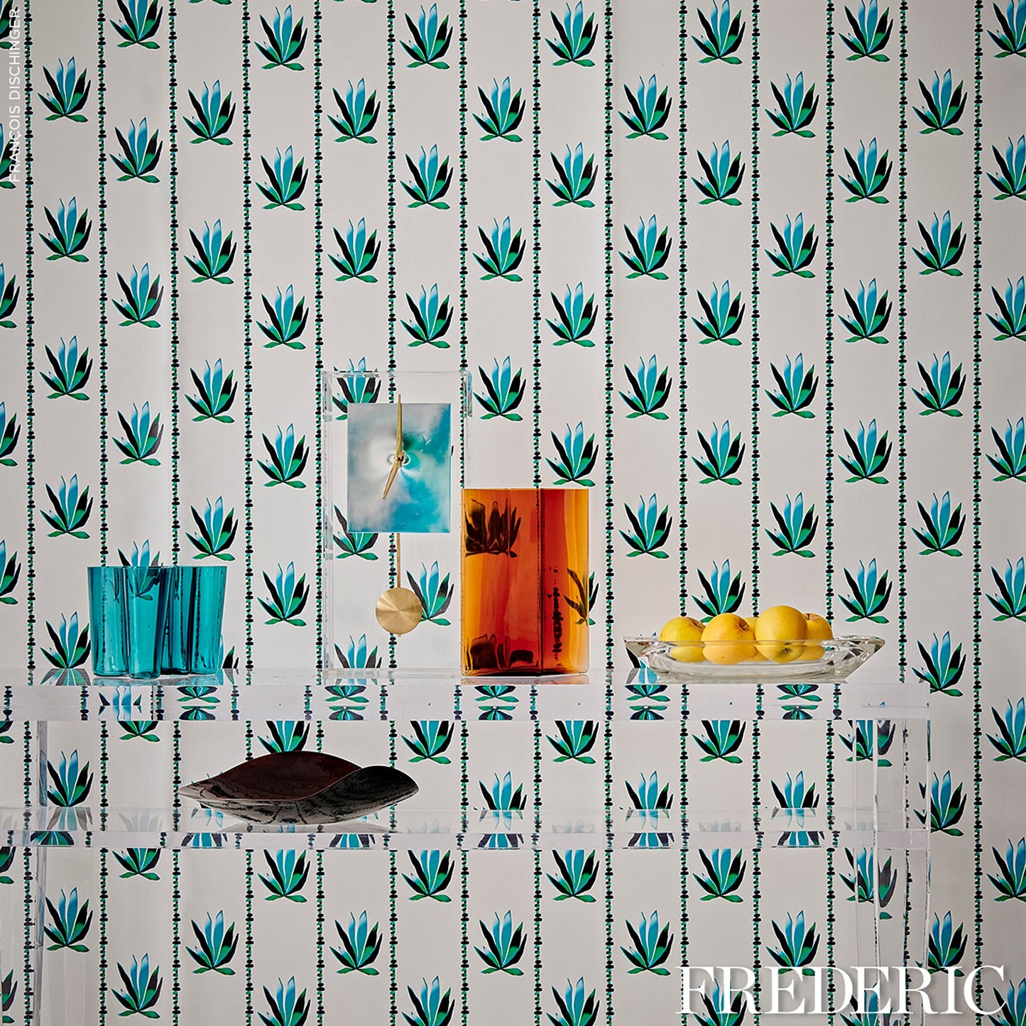 Assortment of colored glass tumblers and vases on teal leaf-pattern wallpaper; plate with lemons.