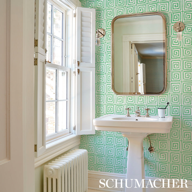 White pedestal sink with brass-framed rounded mirror; teal wallpaper with a leaf pattern; window with shutters and radiator.