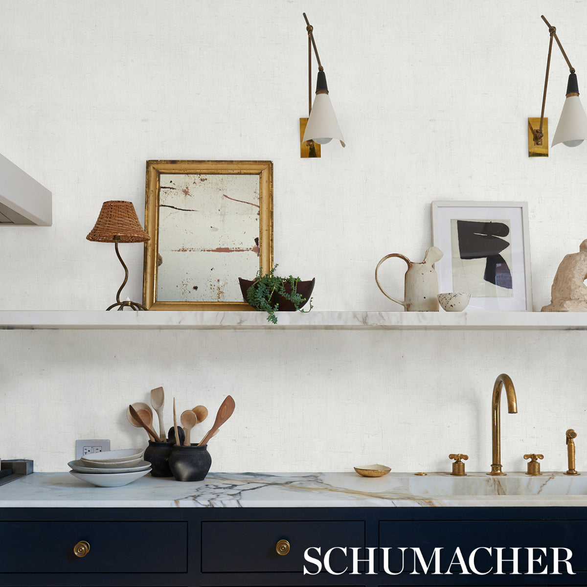 Marble kitchen with navy cabinets, brass faucet, white wall, shelf with art, ceramics, wicker lamp, brass pendant lights.
