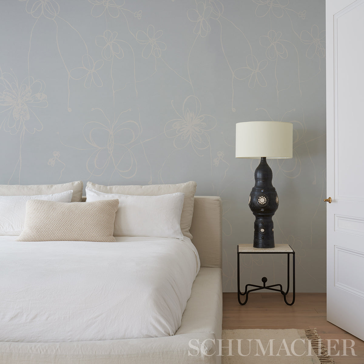 Bedroom with pale blue-gray floral wallpaper, white bedding, beige throw pillow, and a black ornate lamp on a metal-frame nightstand.