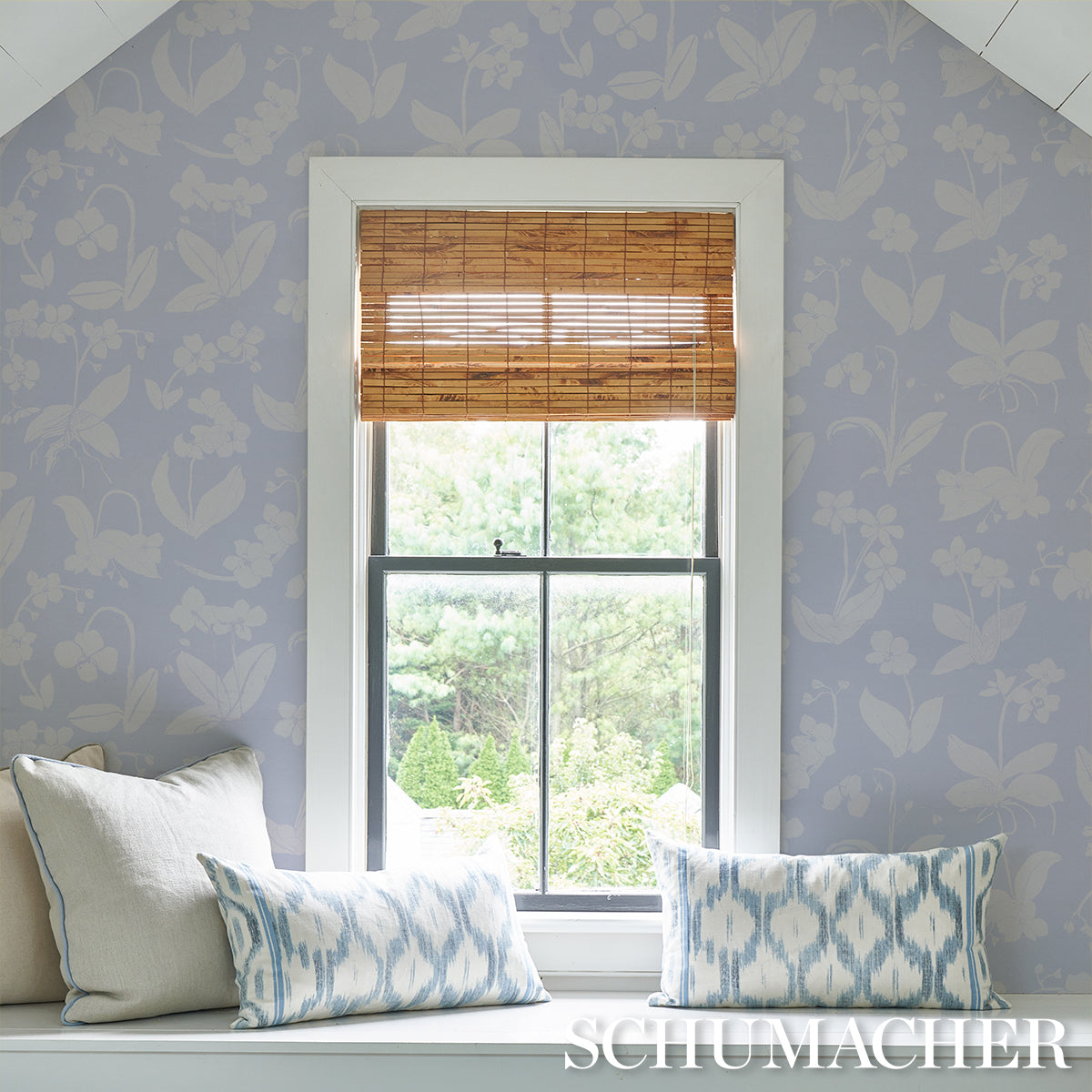 White-framed window with a bamboo shade, lavender-blue floral wallpaper, and a window seat with blue ikat cushions.