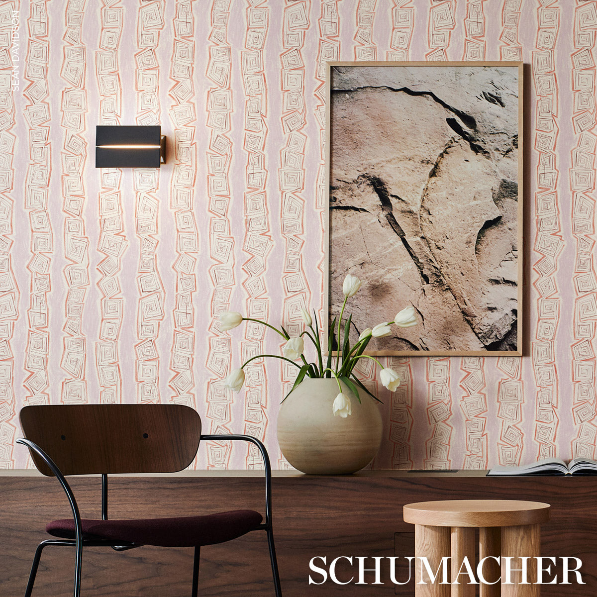 Pink patterned wallpaper, dark wood bench, black metal chair, white tulips in ceramic vase, framed stone art, wood stool.