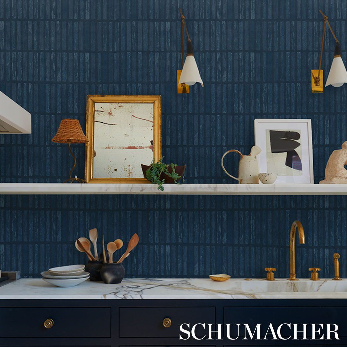 Blue textured kitchen wall with marble countertop, brass faucet, white shelves, frames, and ceramic tableware.