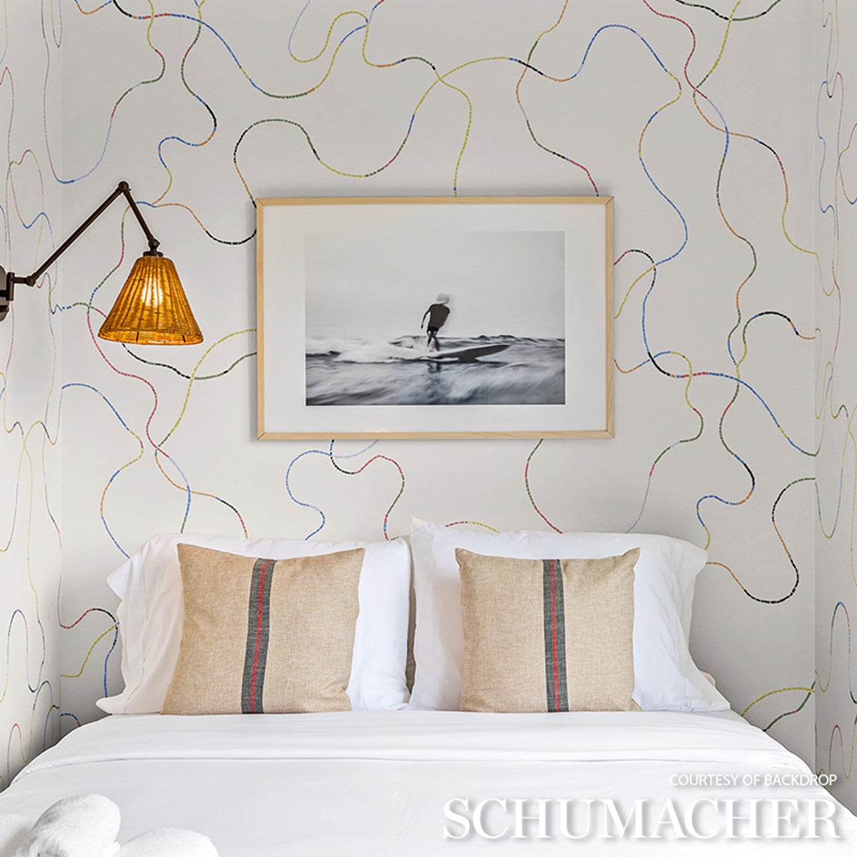 Bedroom with abstract bright multi wallpaper, amber lamp, white bedding and beige striped pillows.