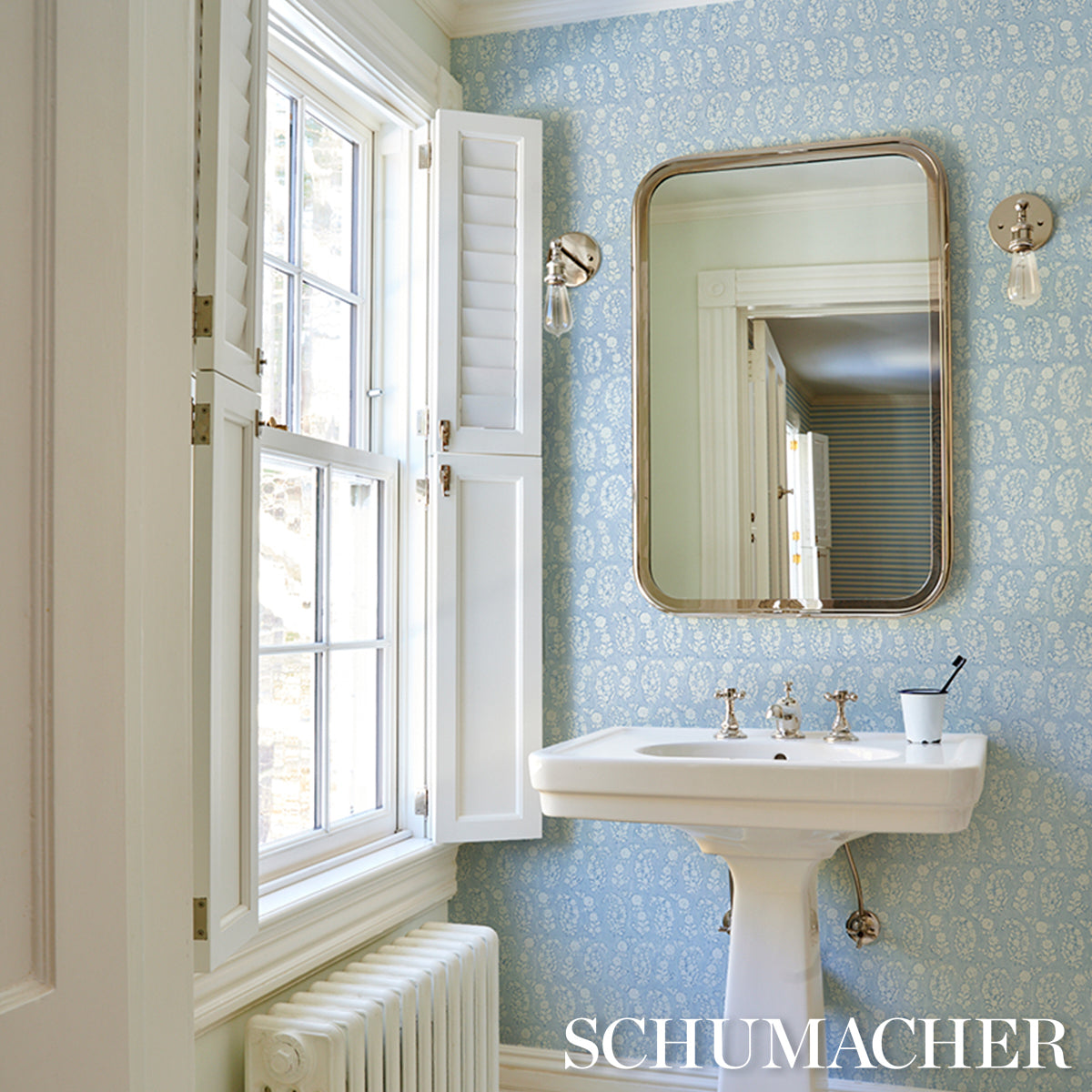 White pedestal sink and rounded mirror set against blue Schumacher damask wallpaper with a sunlit window and wall sconces.