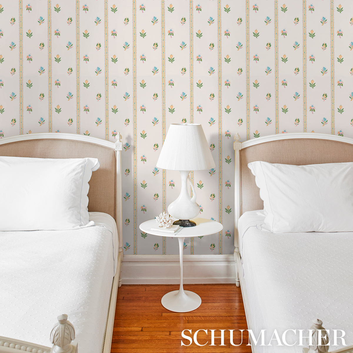 Two twin beds with white bedding and beige headboards, round white nightstand with lamp between them, floral wallpaper.