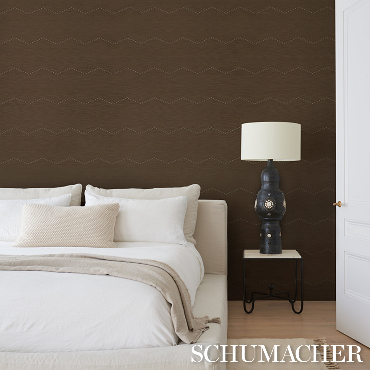 Brown zigzag wallpaper behind a white bed with beige throw and pillows, white shade lamp on a small side table.