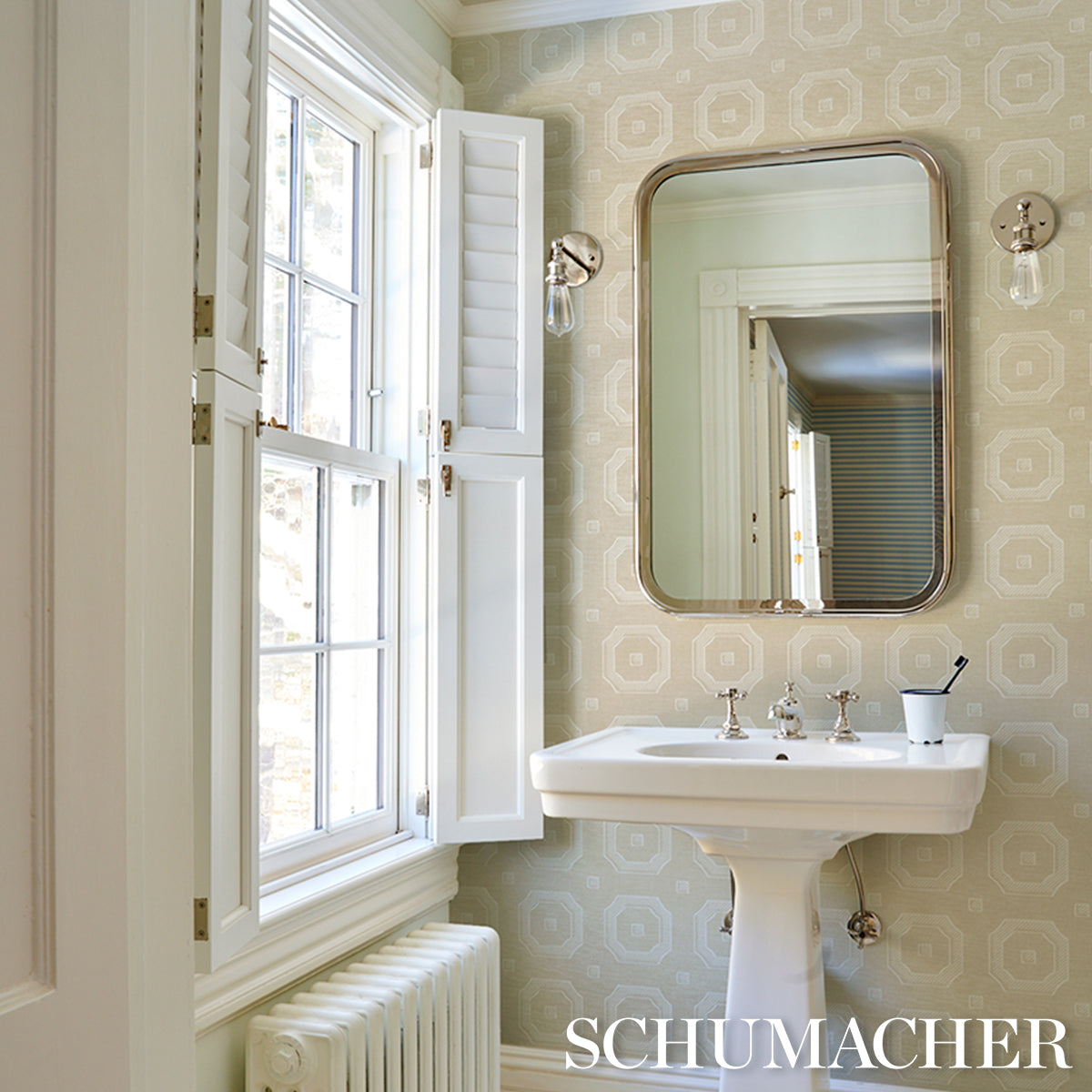 Retro bathroom with pale green Schumacher geometric wallpaper, white pedestal sink, rounded mirror, chrome faucet, window with shutters.
