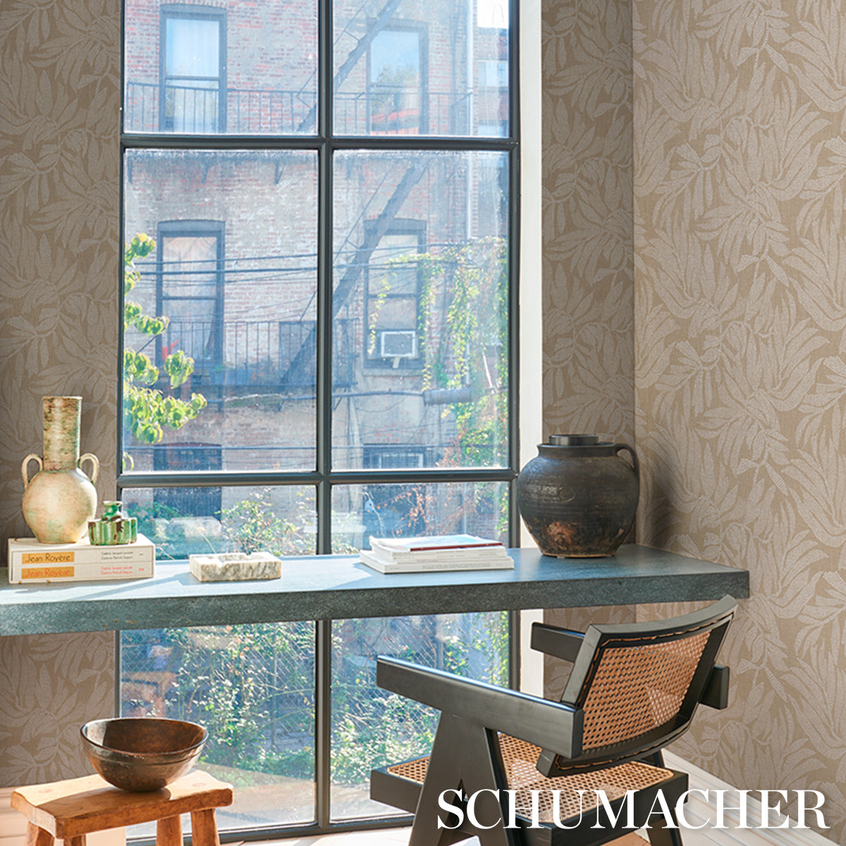 Beige leaf-pattern wallpaper, large grid window, blue-green desk, modern cane-chair, ceramic vase, books.
