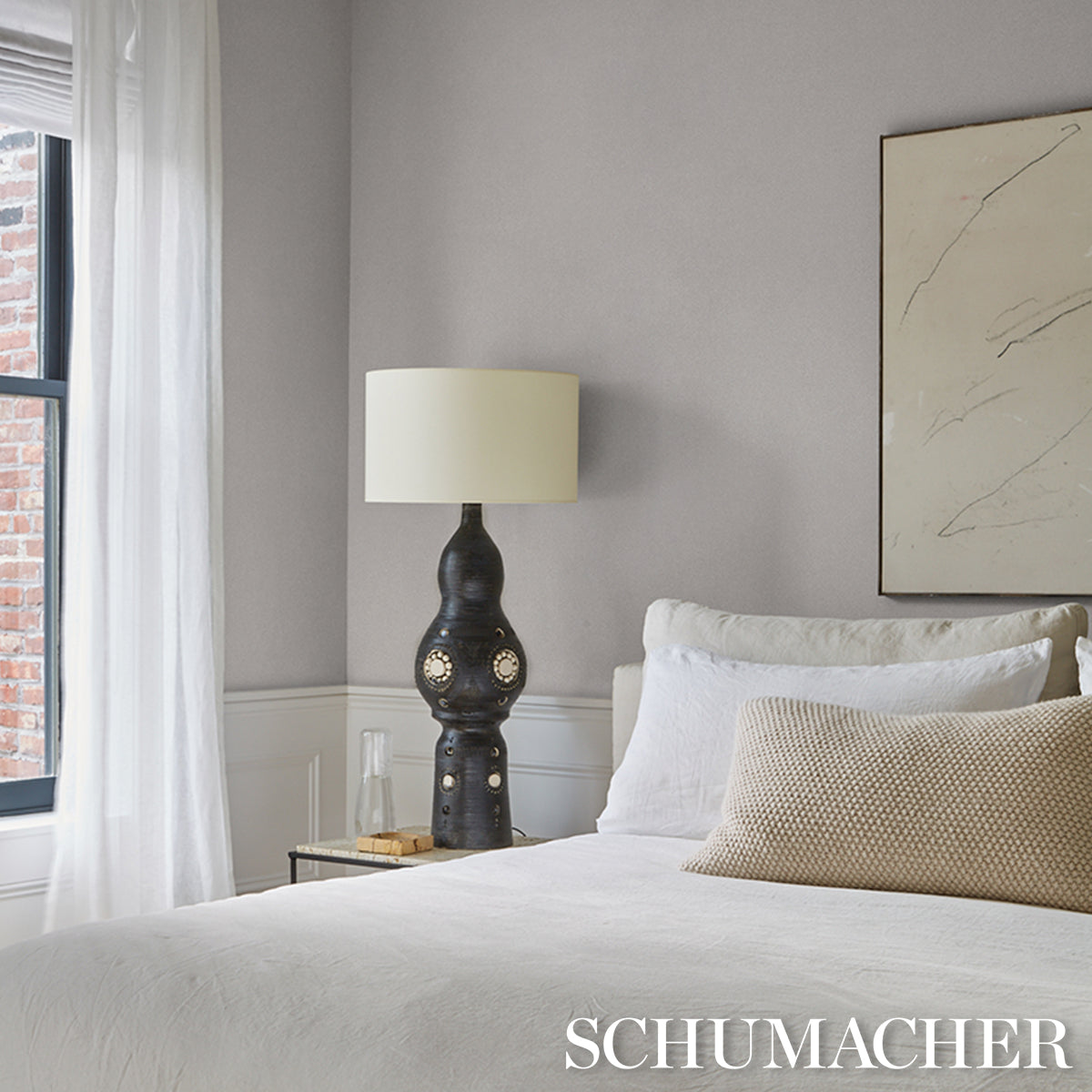 Modern bedroom with a sculptural dark lamp and beige shade, white bedding, beige throw pillow, glass nightstand, marble art.