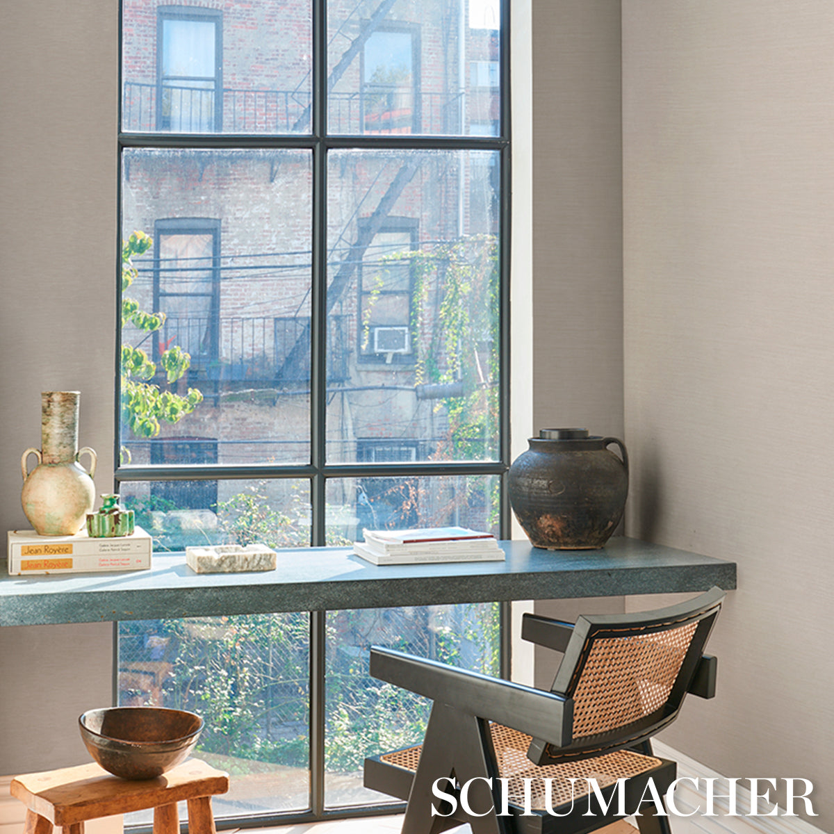 Slate-gray desk by a large grid window; mid-century wicker chair, ceramic jar, brass jug and stacked books.