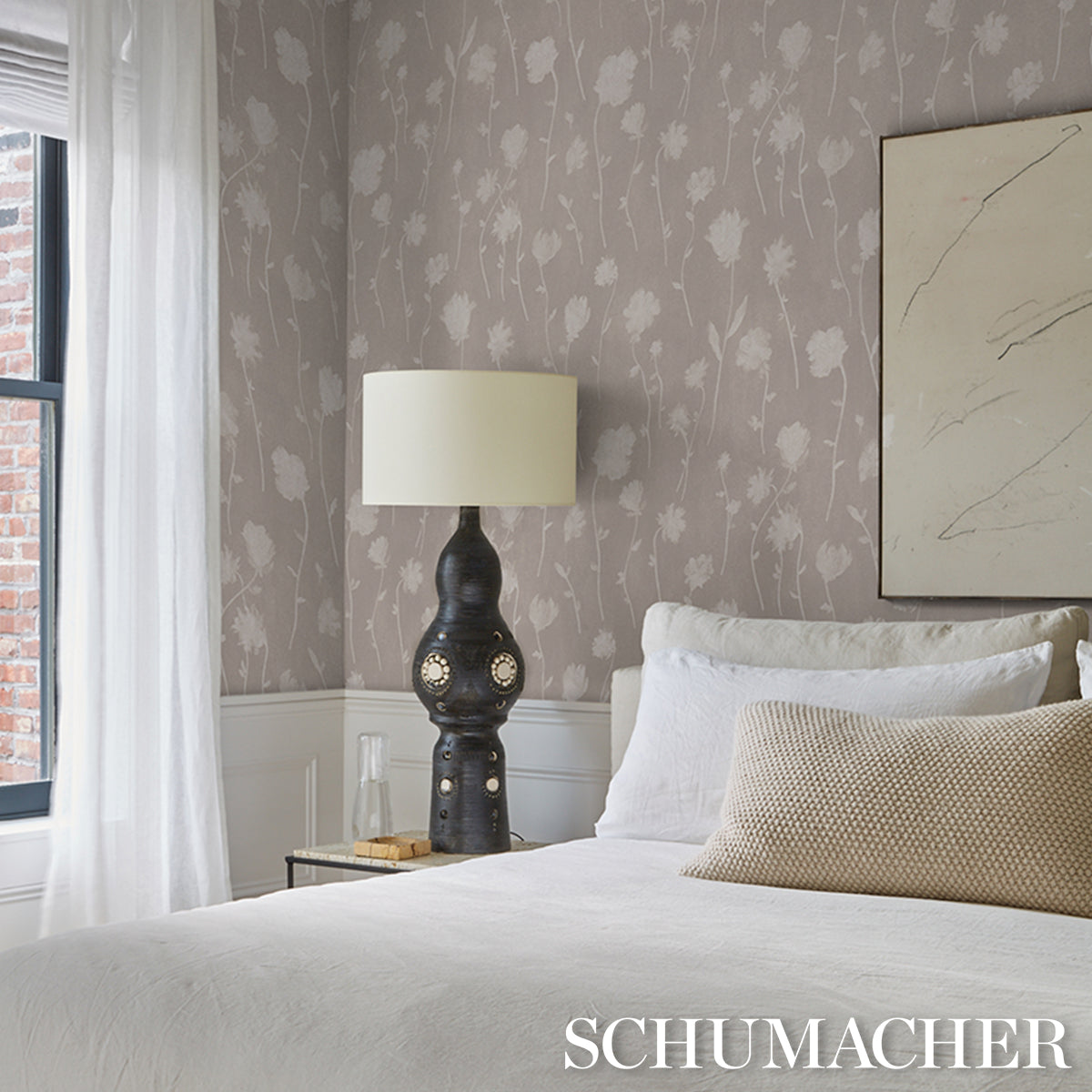 Cozy bedroom with light beige wallpaper, white bedding, beige lumbar pillow; dark sculptural lamp with a cream shade.