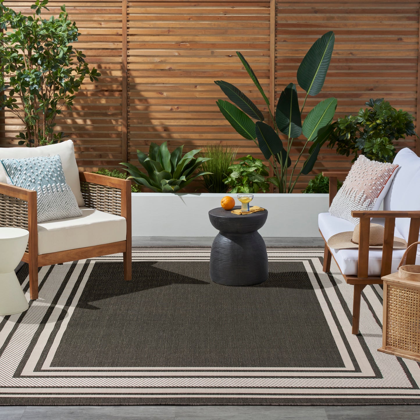 Outdoor patio with beige cushioned wooden chairs, black round table, striped rug, white planter bed, and lush plants.
