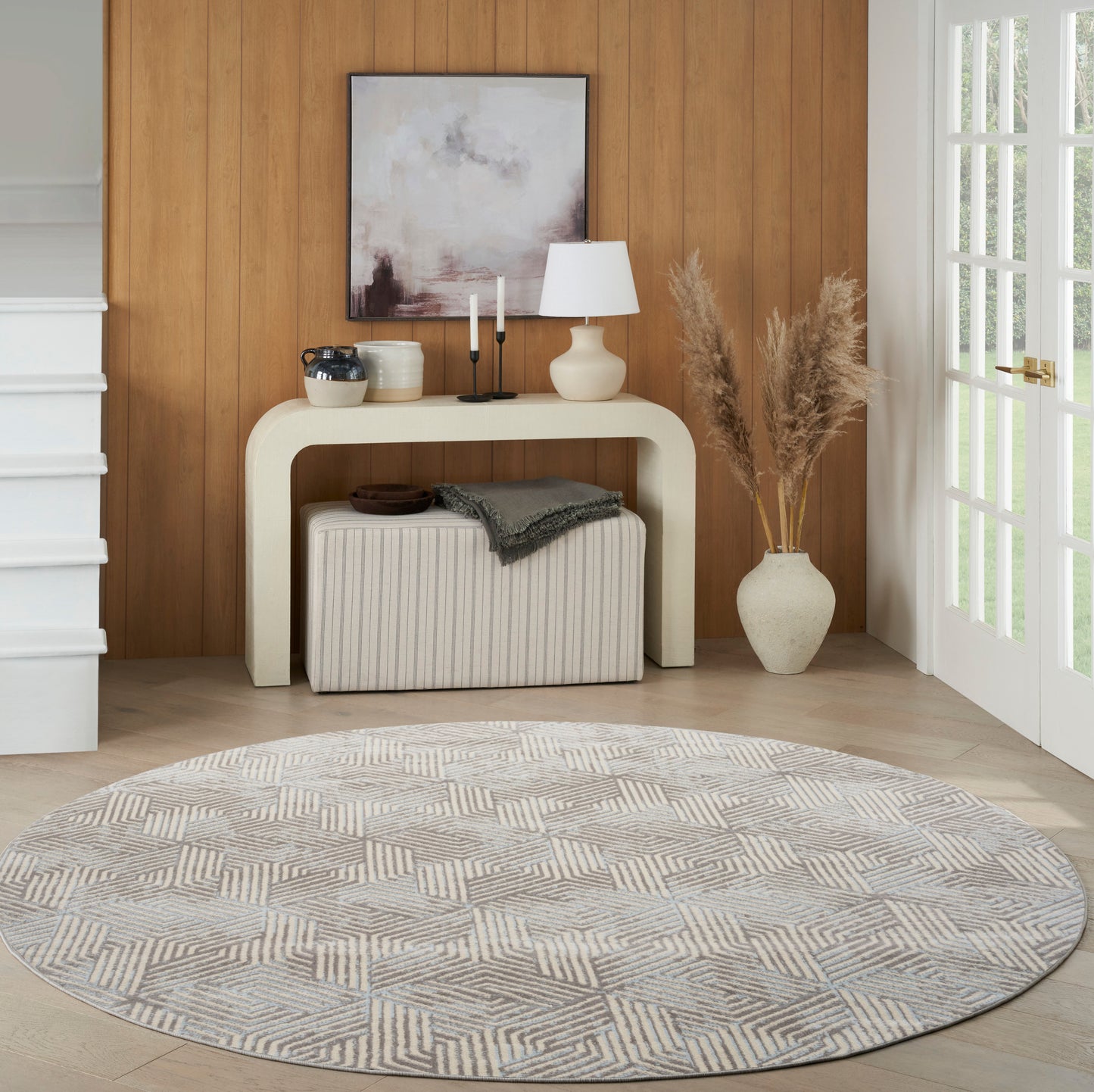 Neutral-toned entryway with a round geometric rug, cream curved console, ottoman, abstract art, lamp, candles, and pampas grass vase.