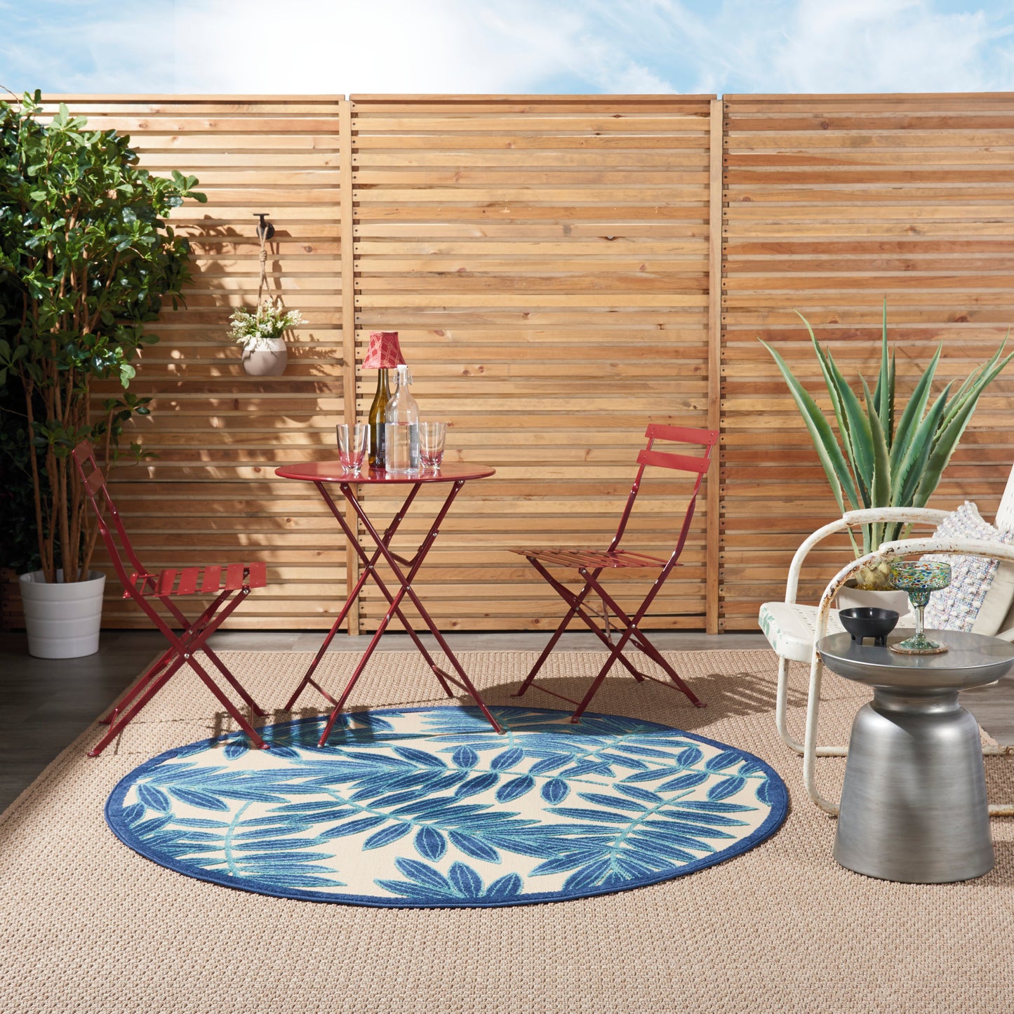 Outdoor red metal bistro set on a round blue palm-leaf rug with potted plants and white wicker chair by a wooden fence.