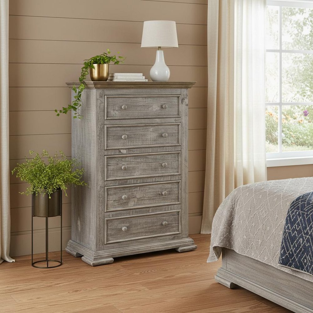Weathered gray five-drawer dresser in a sunlit bedroom, topped with a white lamp, books, and trailing plant.