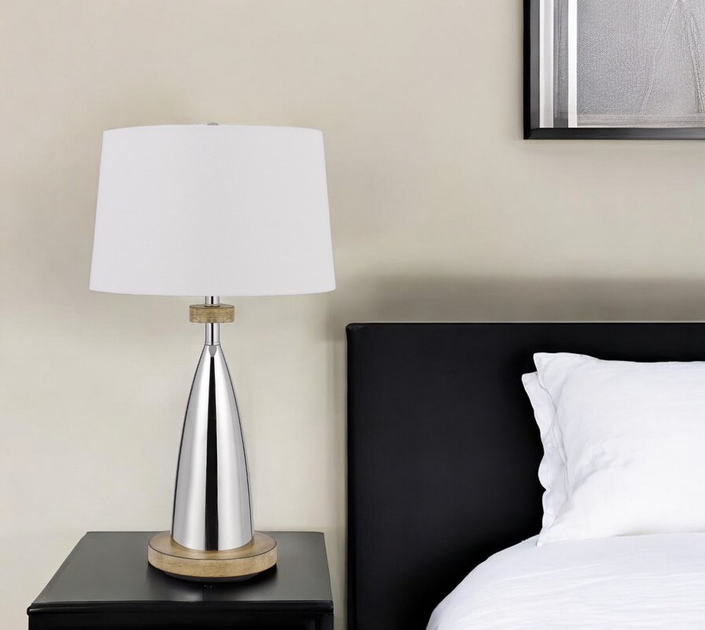 Modern silver cone table lamp with wooden base & white drum shade on a black nightstand beside a white bed.