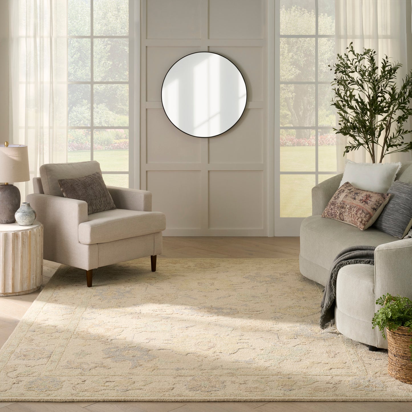 Bright neutral living room with round black-framed mirror, beige armchair, gray sofa, sheer curtains, and cream rug.