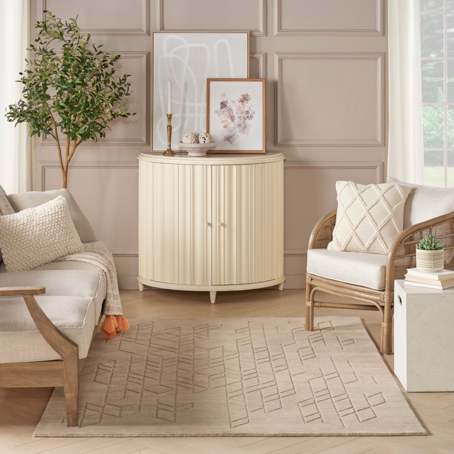 Soft beige living room with curved cream cabinet, framed art, potted plant, rattan armchair, neutral sofa, and rug.