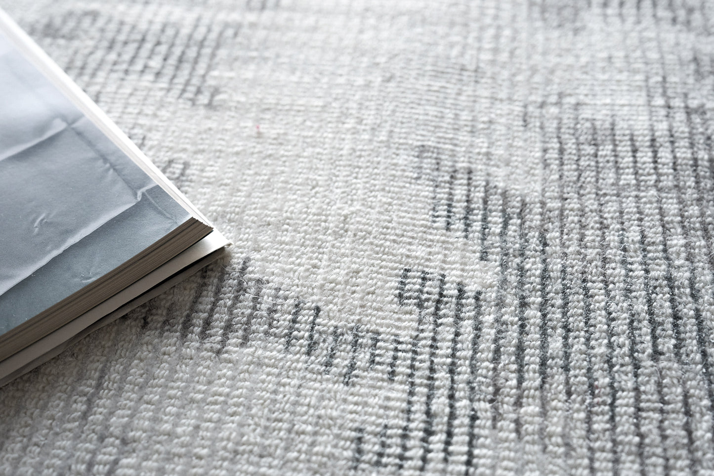 Gray rug with a textured surface and dark linear patterns; an open light gray magazine with white pages rests on it.