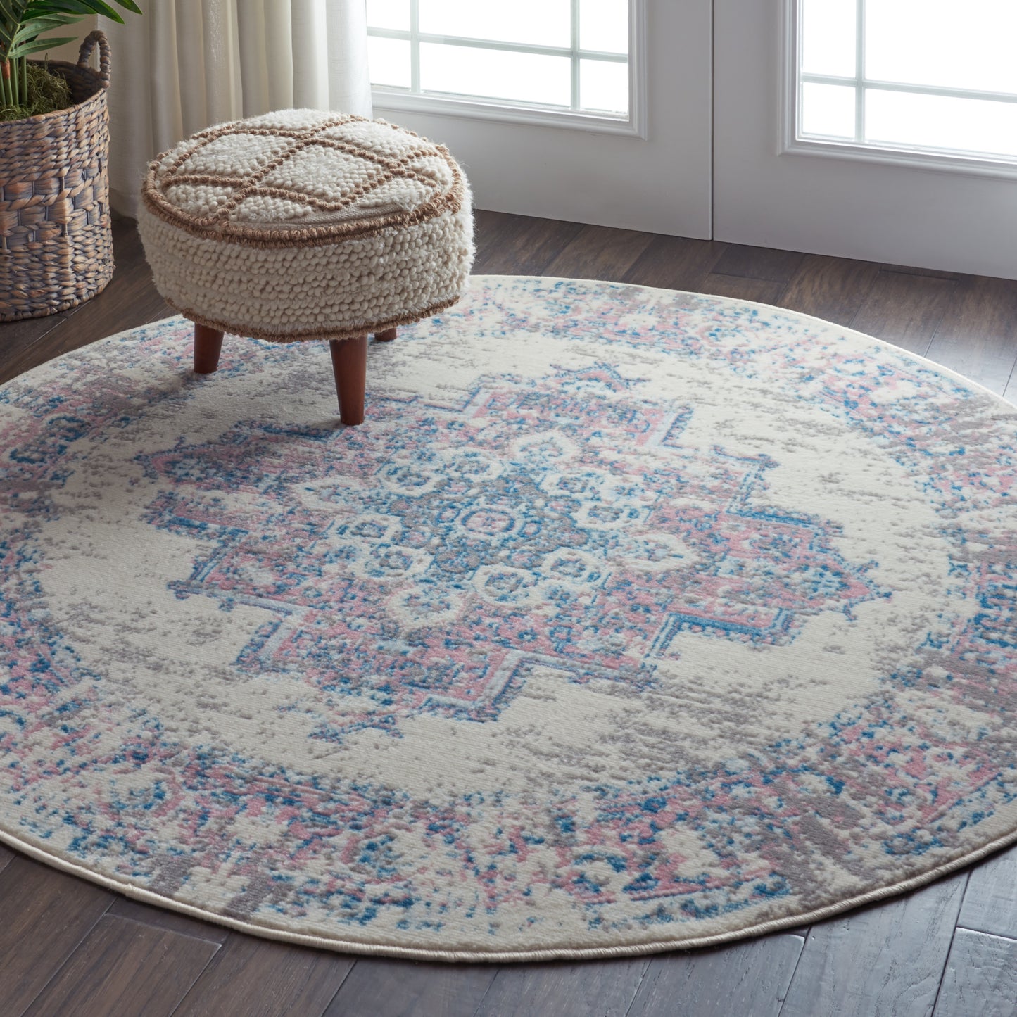 Round pastel geometric area rug in cream, blue and pink with a beige knitted pouf on wooden legs near a plant basket.