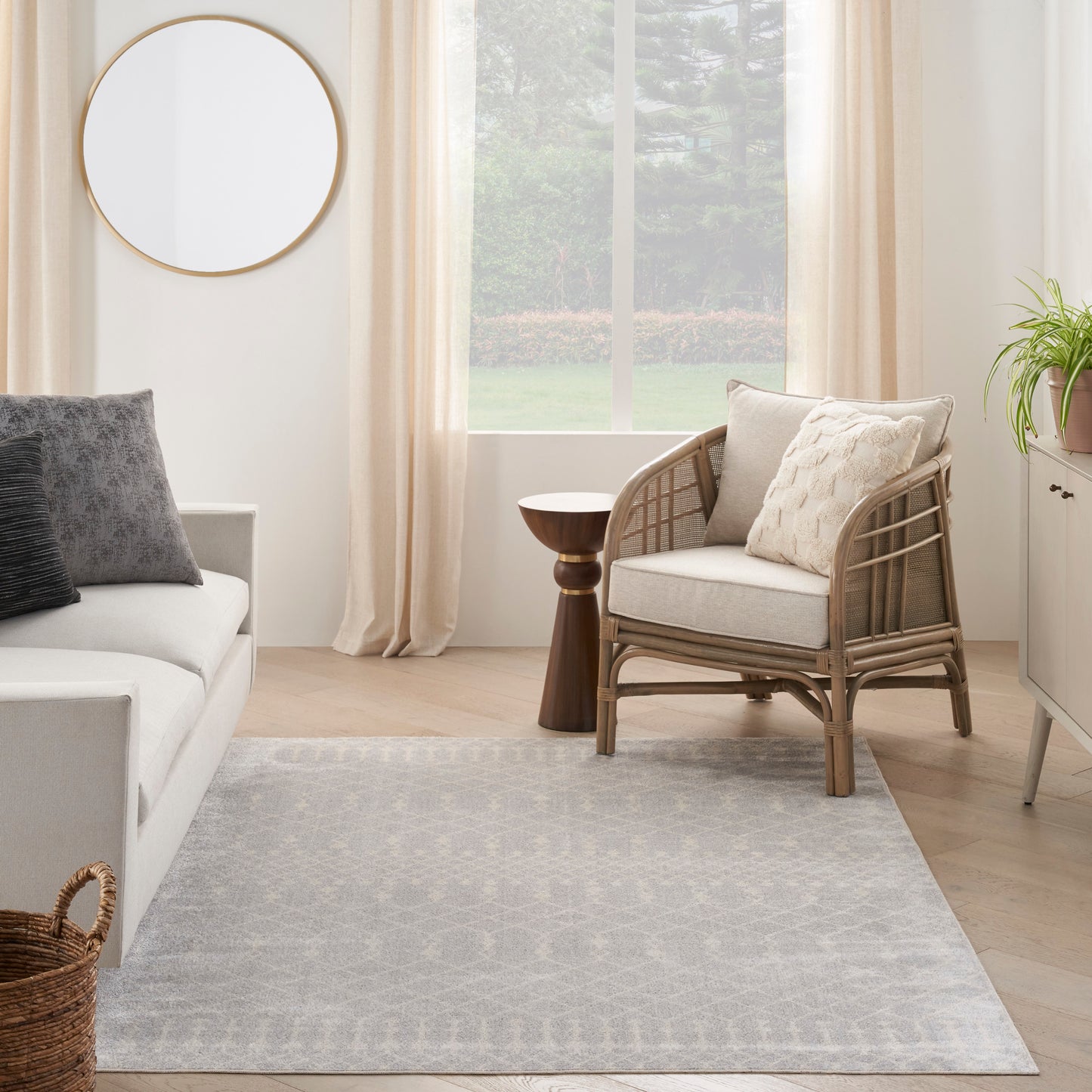 Bright living room with light wood floors, a rattan armchair with beige cushions, circular mirror, and a gray patterned rug.
