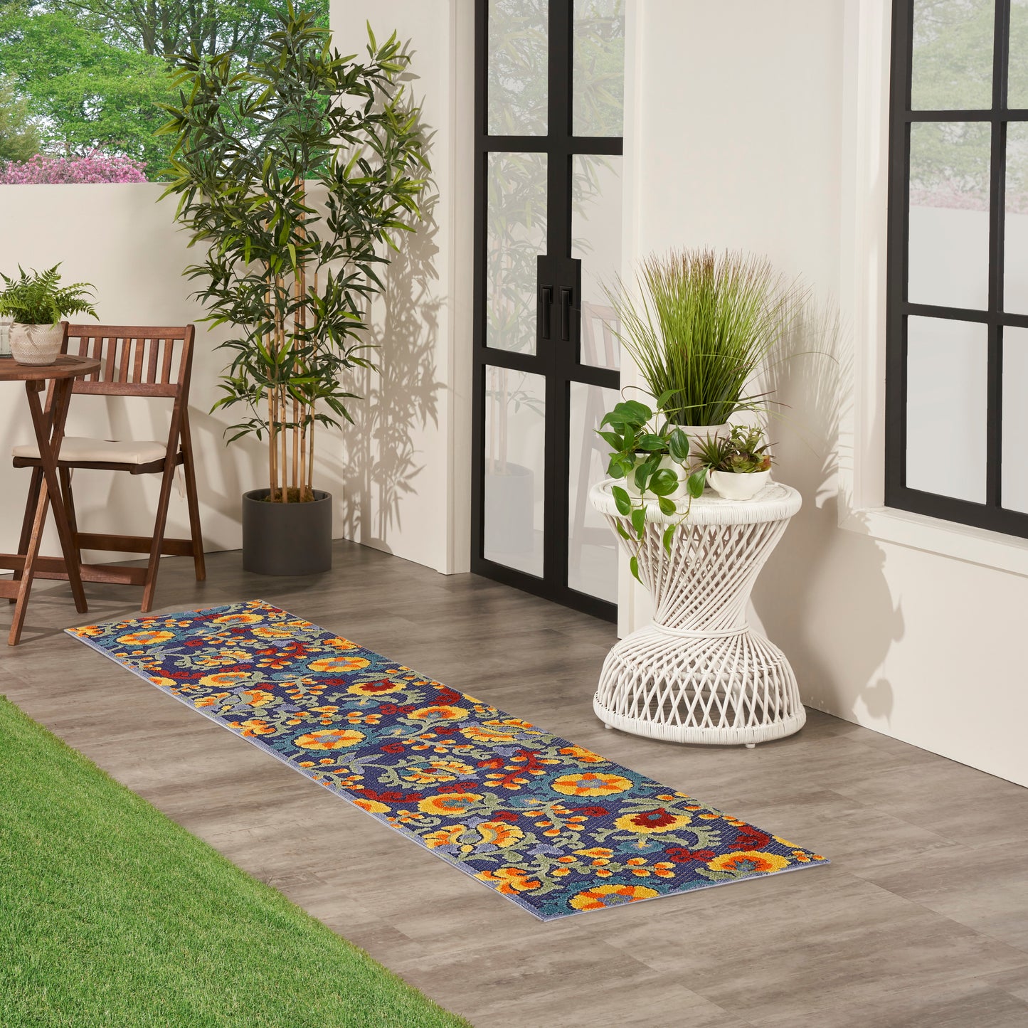 Colorful floral runner rug on a blue background, gray wood floor beside a chair and tall plant, black-framed glass doors.