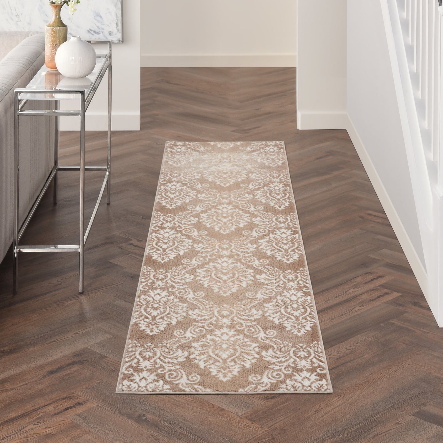Beige damask runner rug on dark wood herringbone floor; glass console table left, white staircase railing right.