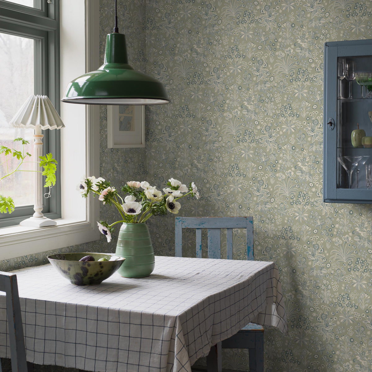 Blue dining nook with checkered tablecloth, green pendant lamp, ceramic vase of white flowers, blue chairs, floral wallpaper