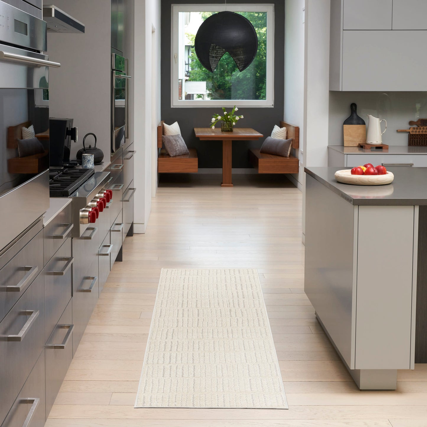 Modern kitchen with white island, light wood floors, beige runner, and dining nook with wooden benches under a black pendant.