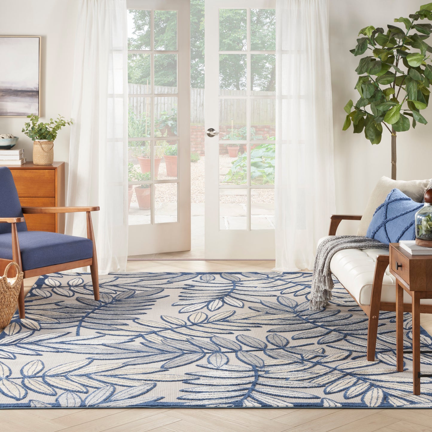 Bright living room with white sheer curtains, blue leaf rug, wooden coffee table, sofa with cushions, and a potted plant.