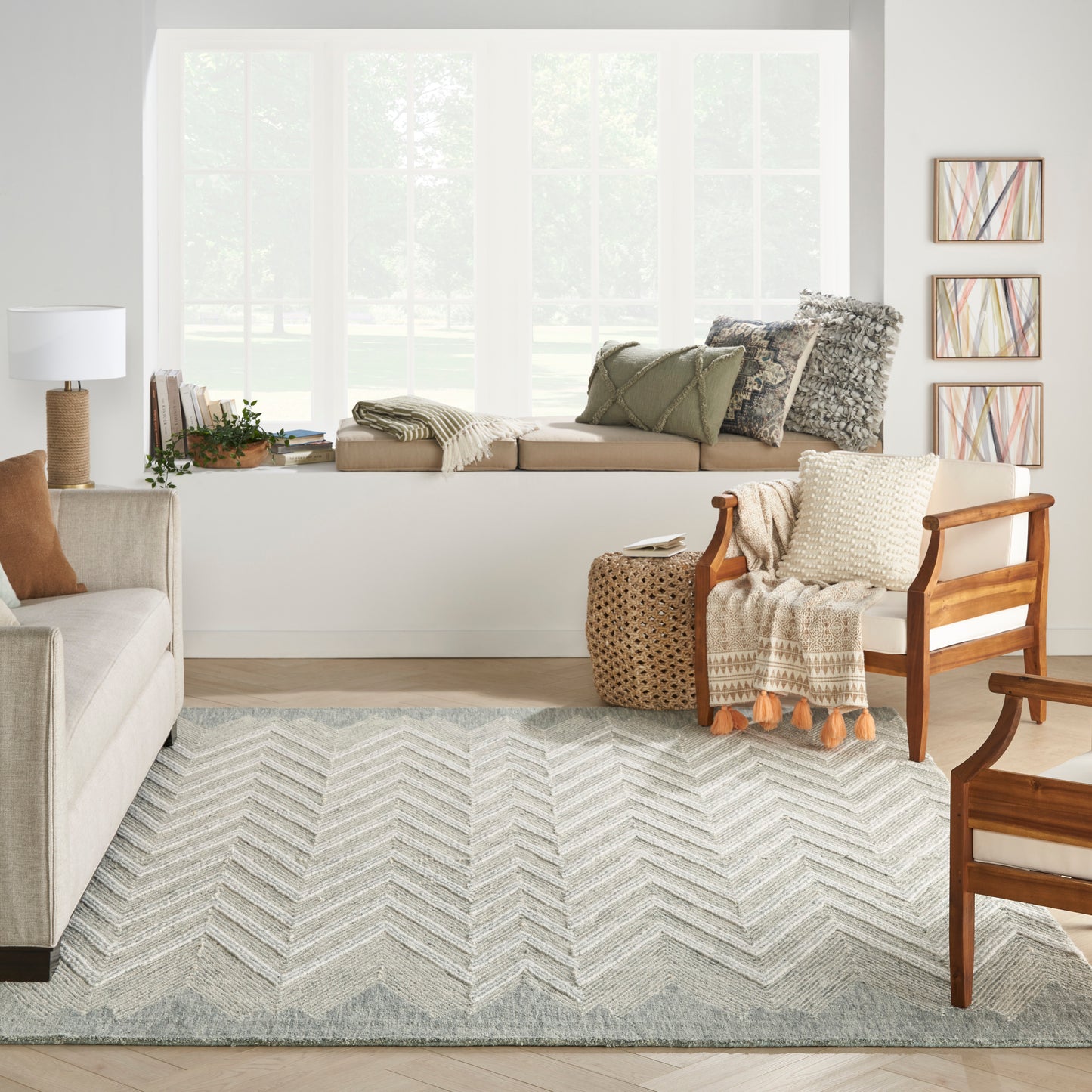 Cozy living room with window seat, beige sofa, wooden chairs, chevron rug, knit throw, wicker table, and abstract art.