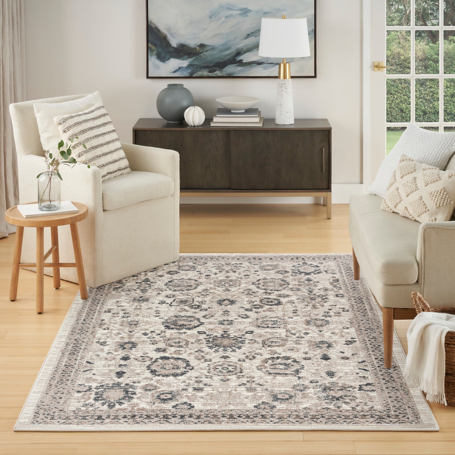 Cozy living room with beige seating, ornate gray rug, wooden side table with plant, dark credenza, abstract art, and lamp.