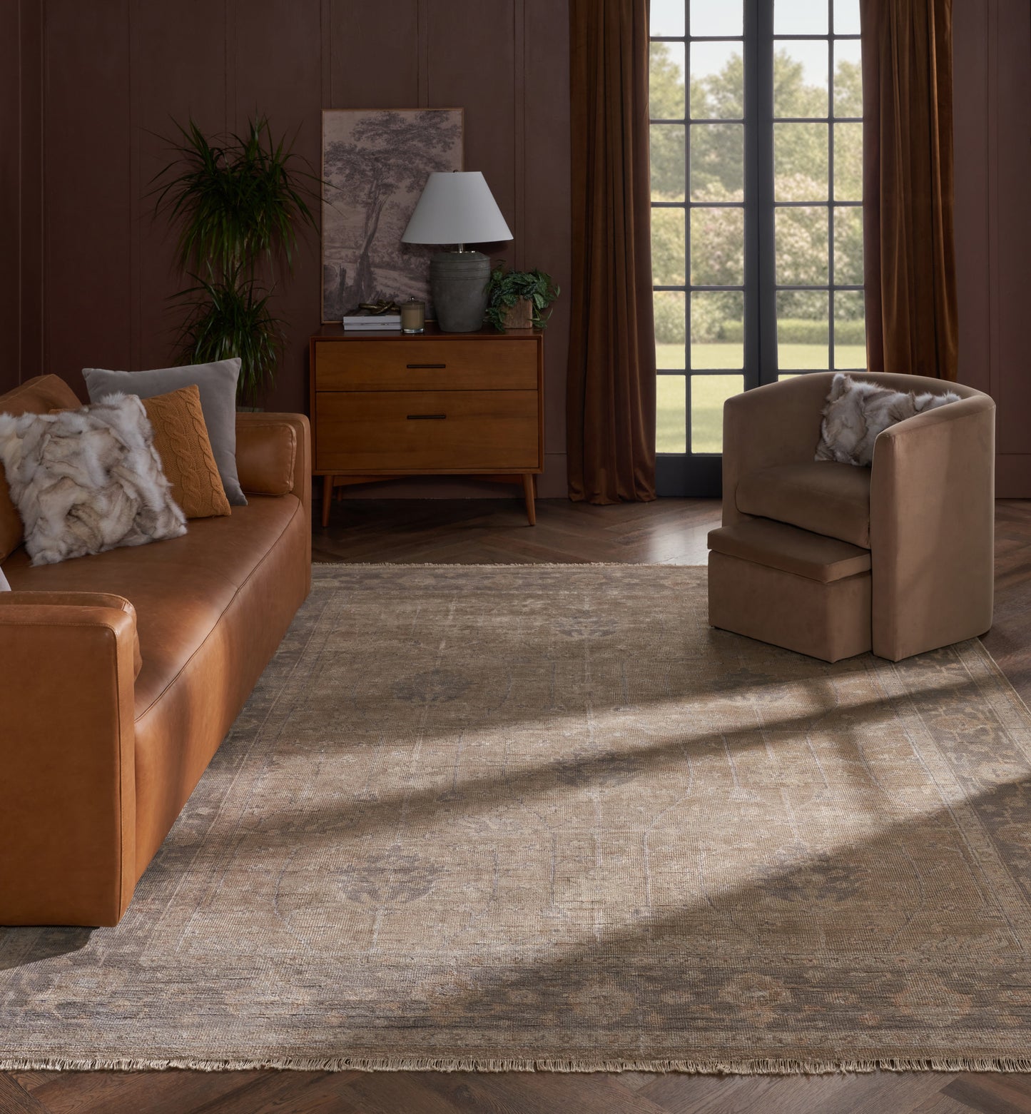 Warm living room with tan leather sofa, matching armchair, wooden dresser, lamp, plants, and a muted patterned area rug.