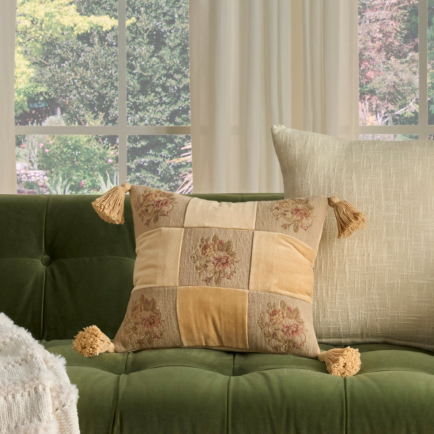 Floral patchwork cushion with tassels on a green tufted sofa by a window