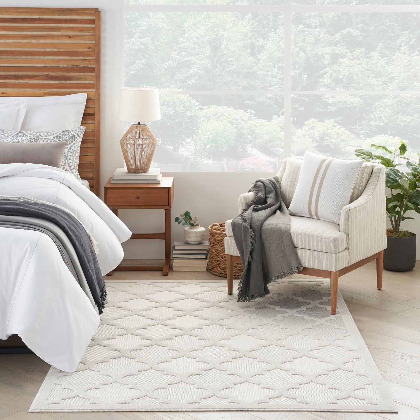 Bright modern bedroom with wooden headboard, white bedding, cream armchair, gray throw, geometric area rug, and greenery.