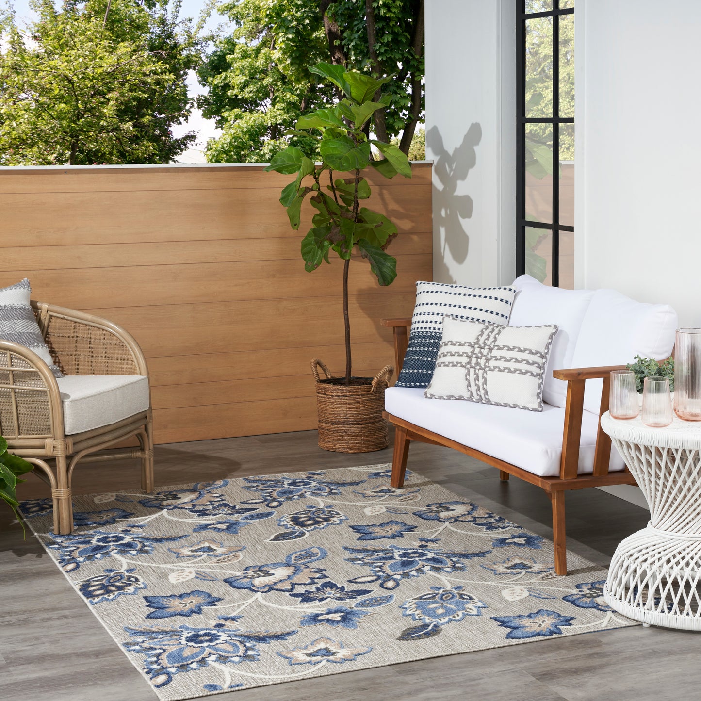 Outdoor balcony with white cushioned wooden sofa, wicker chair, round woven table, potted plant, and a 5' x 8' blue floral rug.