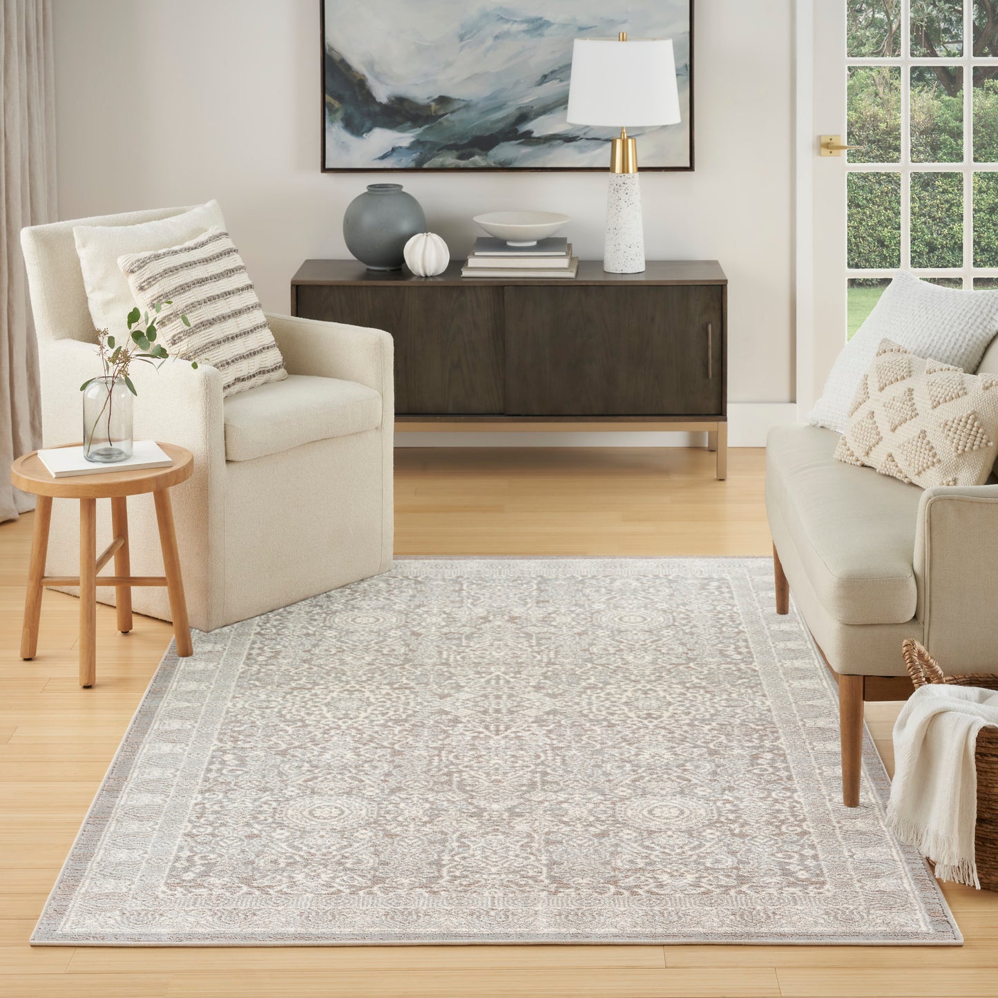 Neutral living room with cream sofa and armchair, round wood side table, gray-cream rug, dark wood credenza, wall art.