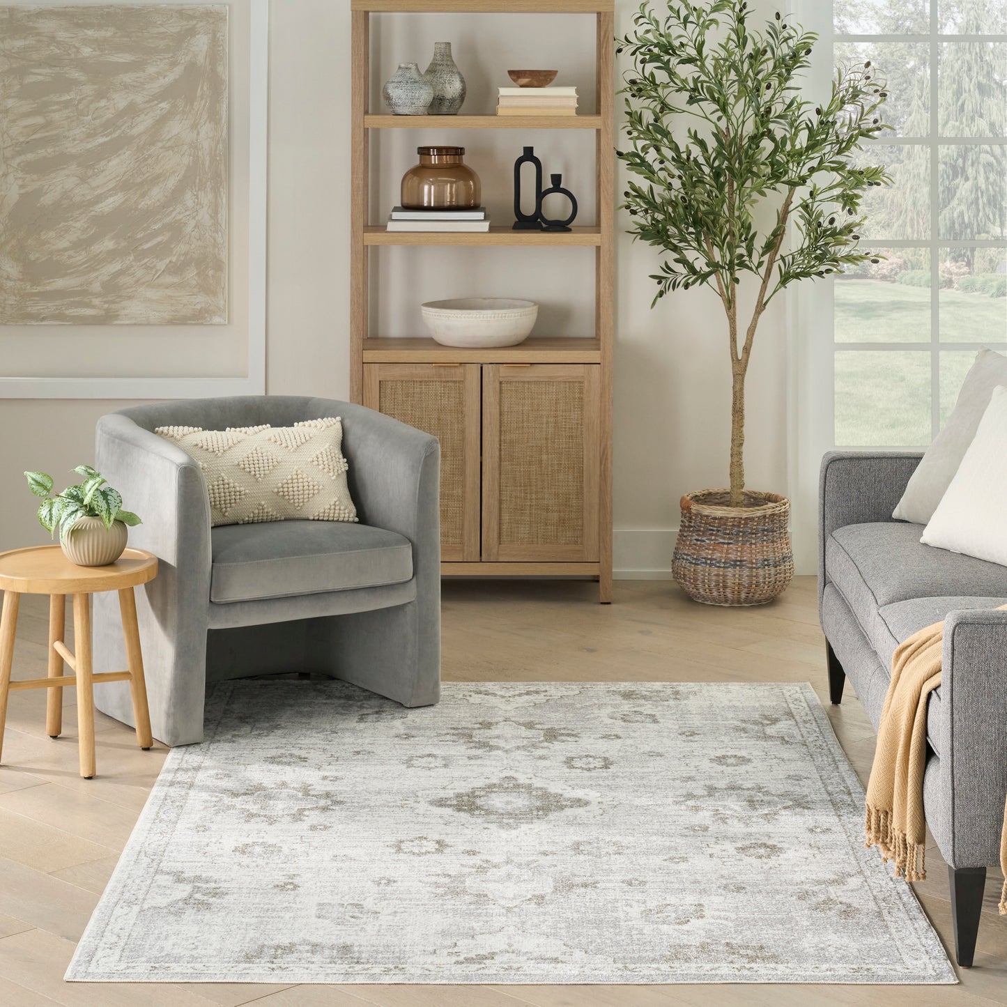 Neutral living room with light-wood shelving, grey upholstered chairs, tall potted tree, and pale patterned grey area rug.