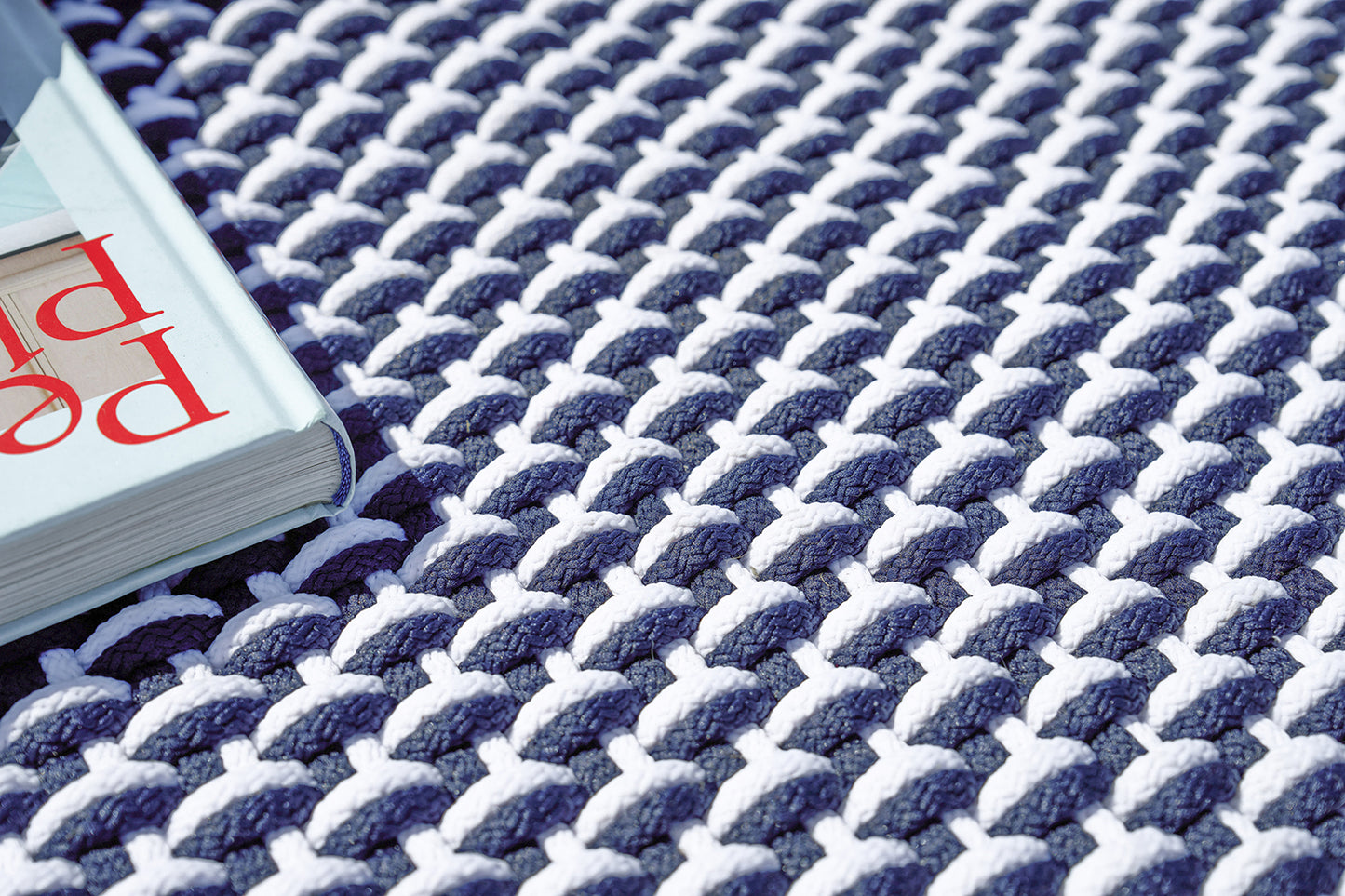 Close-up of navy and white chunky knit rug with thick braided stitches; soft woven texture and a light blue book at edge.