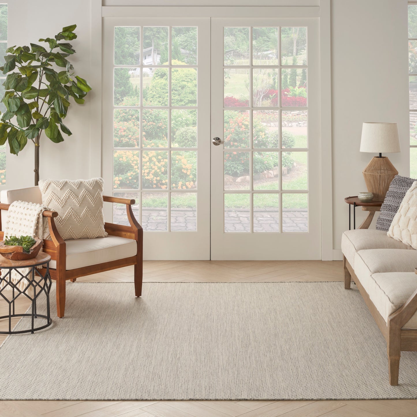 Bright living room with cream sofa, wooden-frame chairs, beige area rug, potted plant, and glass French doors to garden.