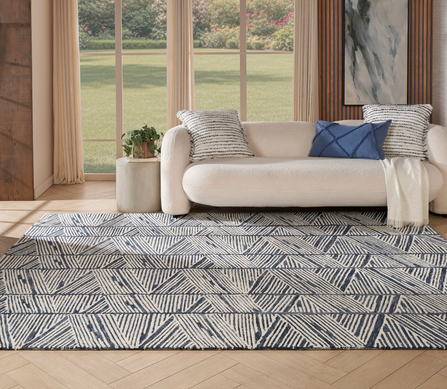 Blue geometric rug on a light wood floor, with beige seating and striped navy cushions draped with a throw, near large windows.