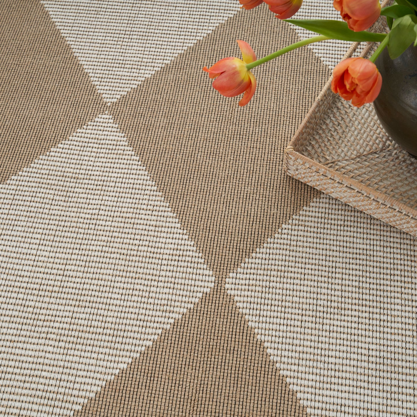 Beige and brown woven rug with large X pattern, beside a wicker tray and orange tulips in a dark vase.