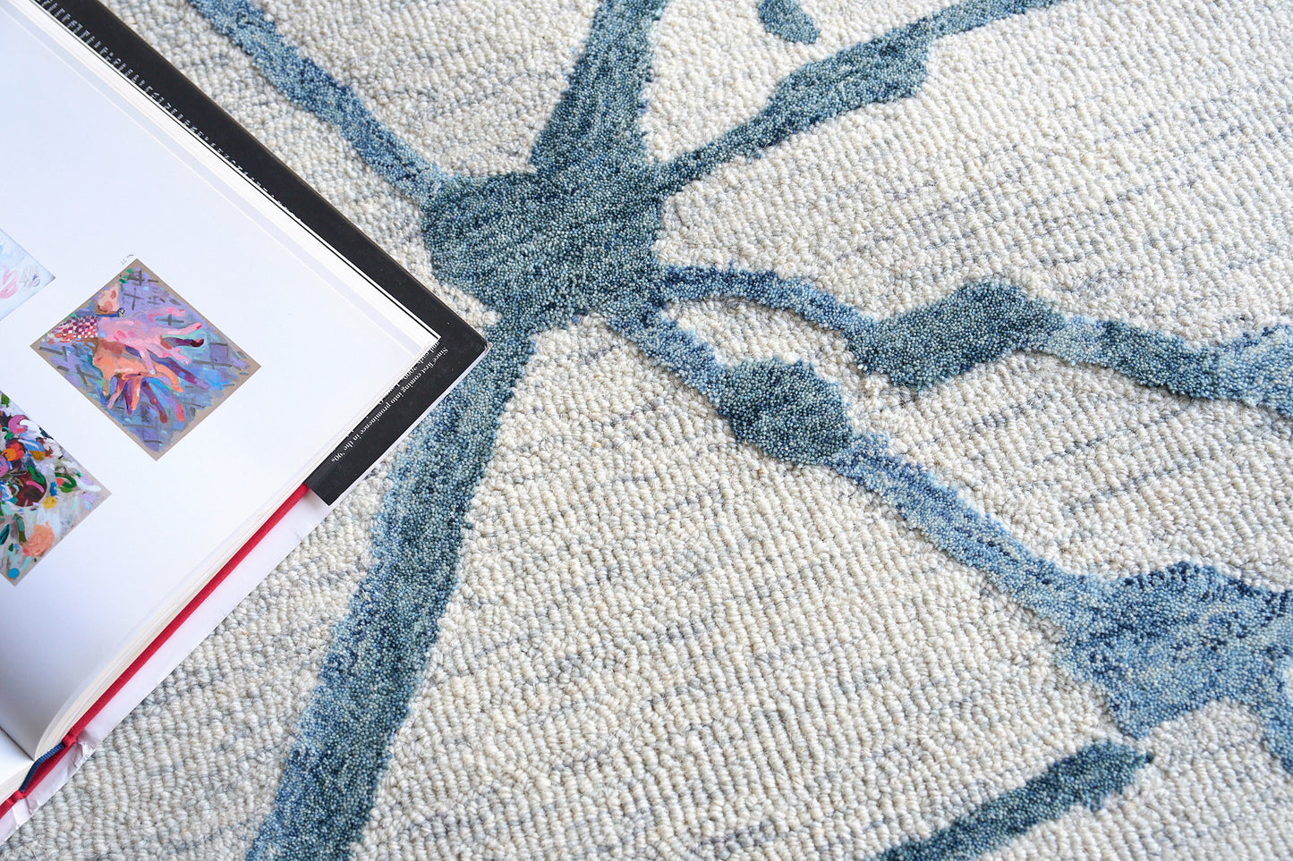 Open art book on a textured cream rug with blue abstract lines and small colorful images.