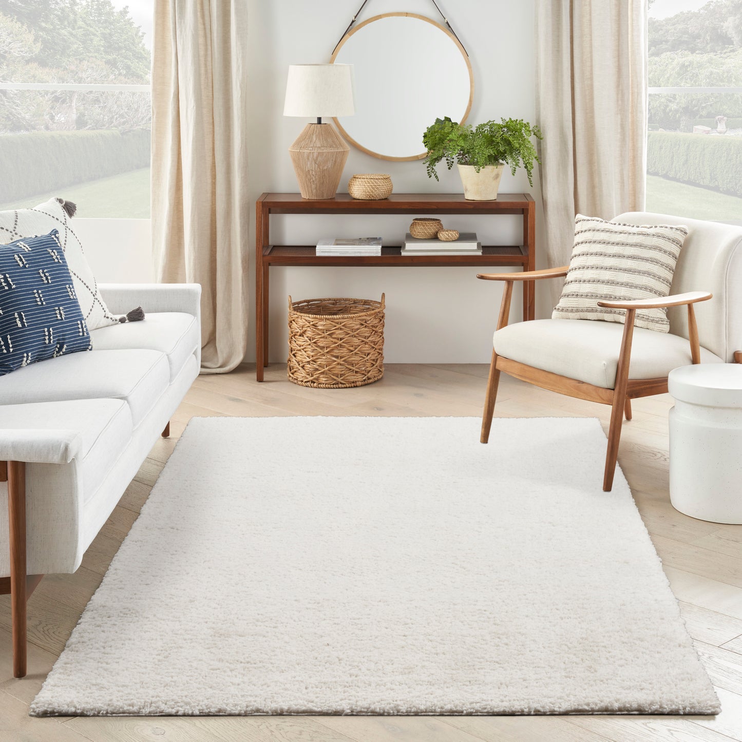 Cream textured area rug anchors a bright living room with mid-century wood chair, white sofa, round mirror, potted plant, and woven basket.