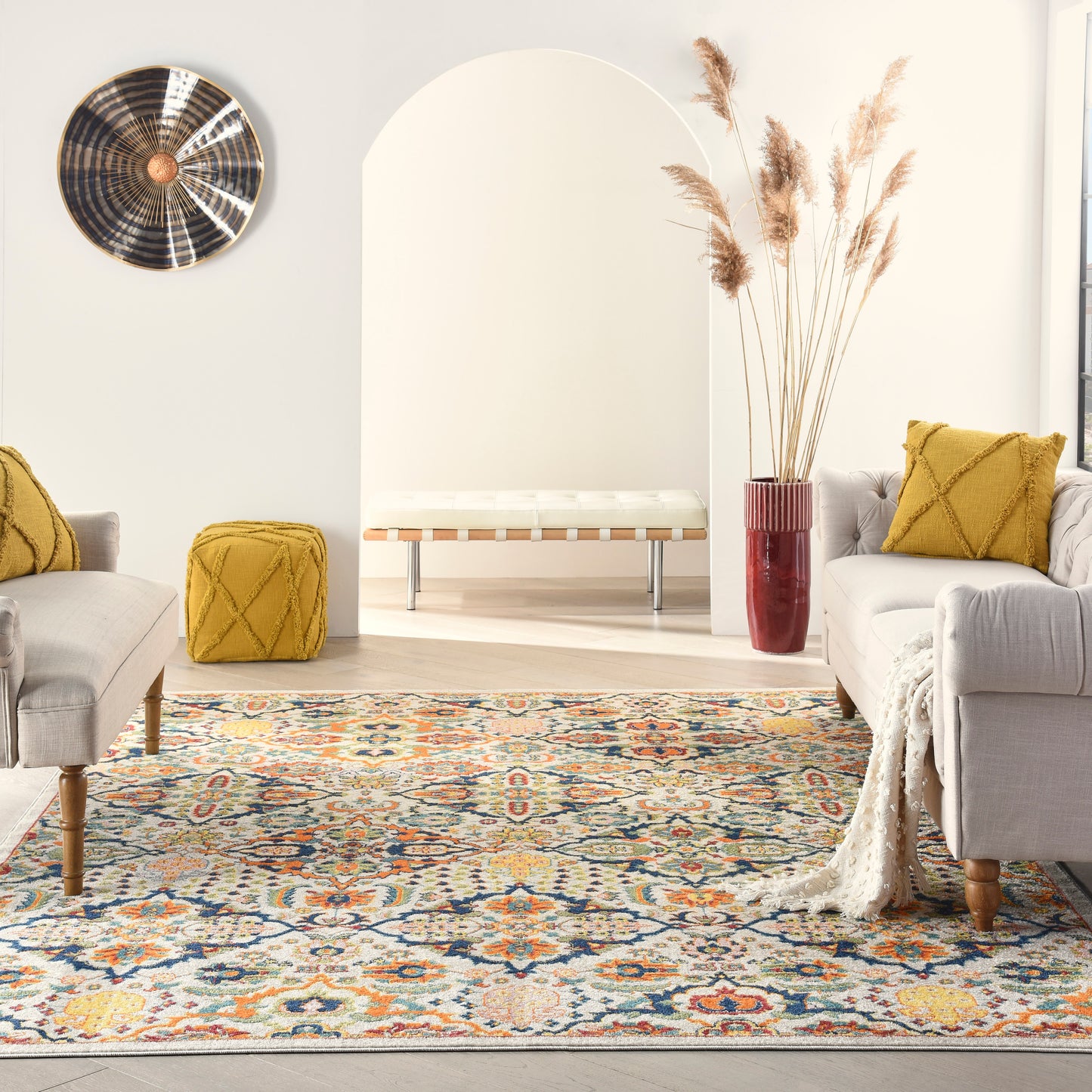 Living room with a vibrant rug, beige sofas, yellow cushions, a tufted yellow ottoman, and a red vase with pampas grass.
