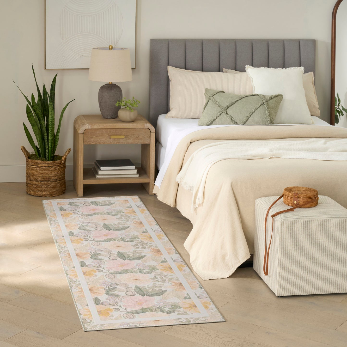 Neutral bedroom with gray tufted headboard, beige bedding, pastel floral rug runner, wooden nightstand, potted plant.