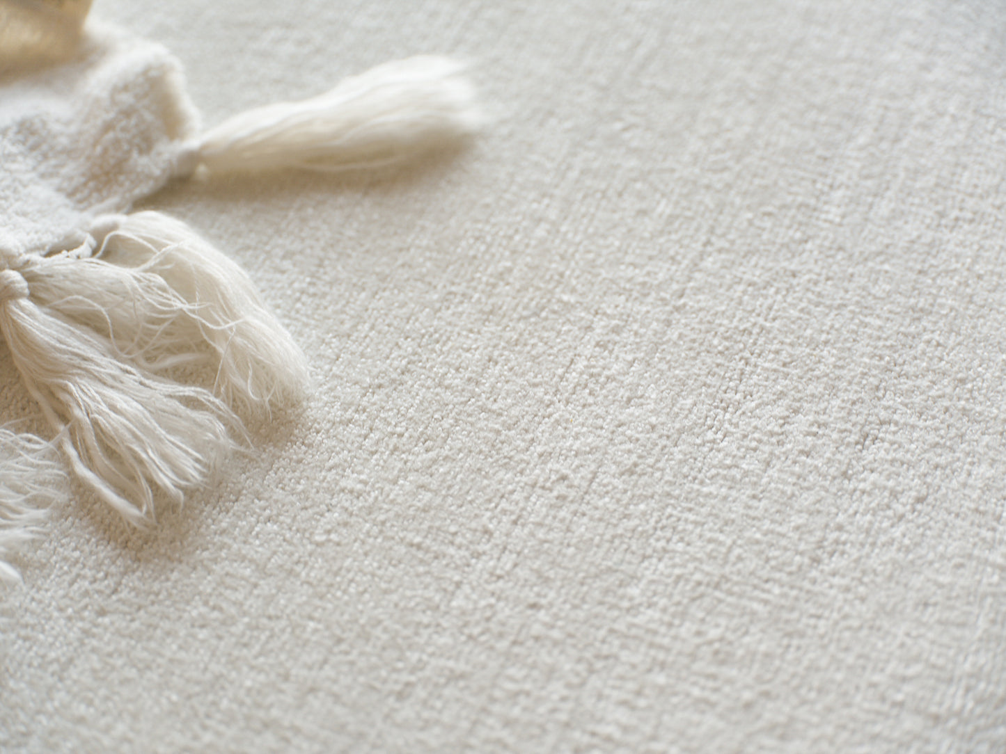 Cream woven throw blanket with tassels on textured beige surface.