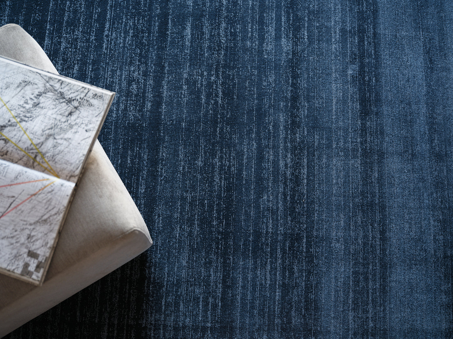 Beige fabric ottoman with an open map spread on top, resting on a dark blue textured rug.