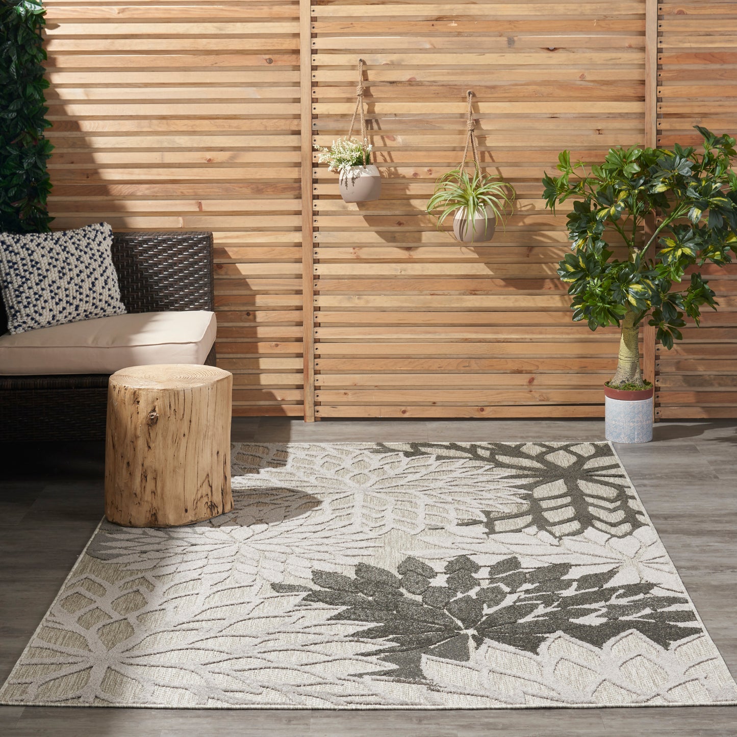 Outdoor patio with wicker sofa, beige cushions, round wood stump table, leaf-pattern rug, hanging planters, potted tree.